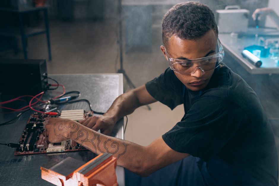 Técnico joven con tatuajes y gafas de protección trabajando en electrónica dentro del taller